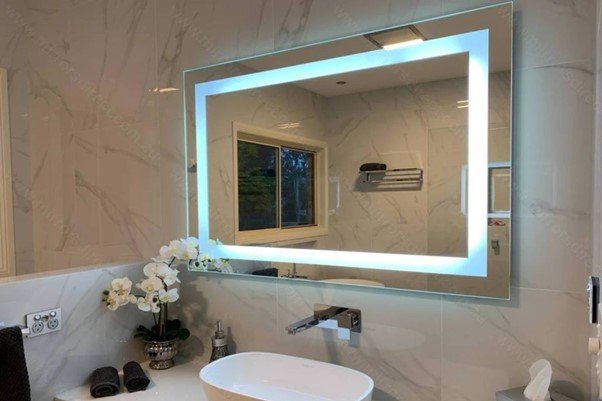 a bathroom mirror decorated by LED lights with sink, flowers, hand towels are on the cabinet