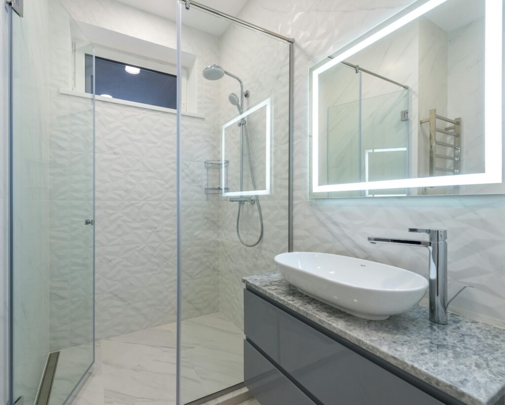 Stylish bathroom mirror shining with LED lights in bathroom above sink with faucet and shower cabin