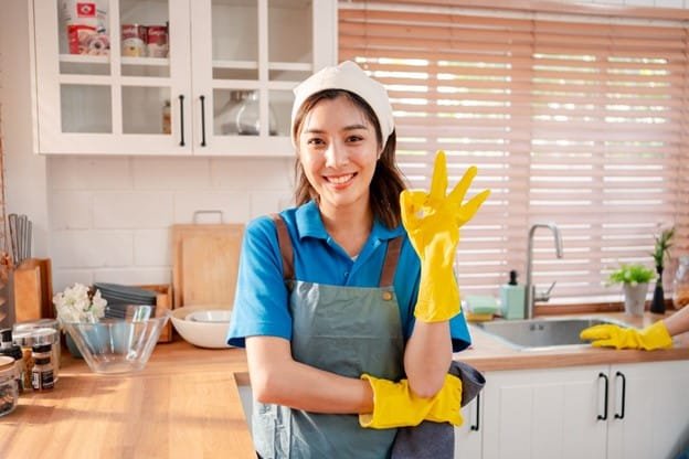 a woman in the picture where she wears a yellow gloves, blue shirt and standing in her kitchen for cleaning the kitchen