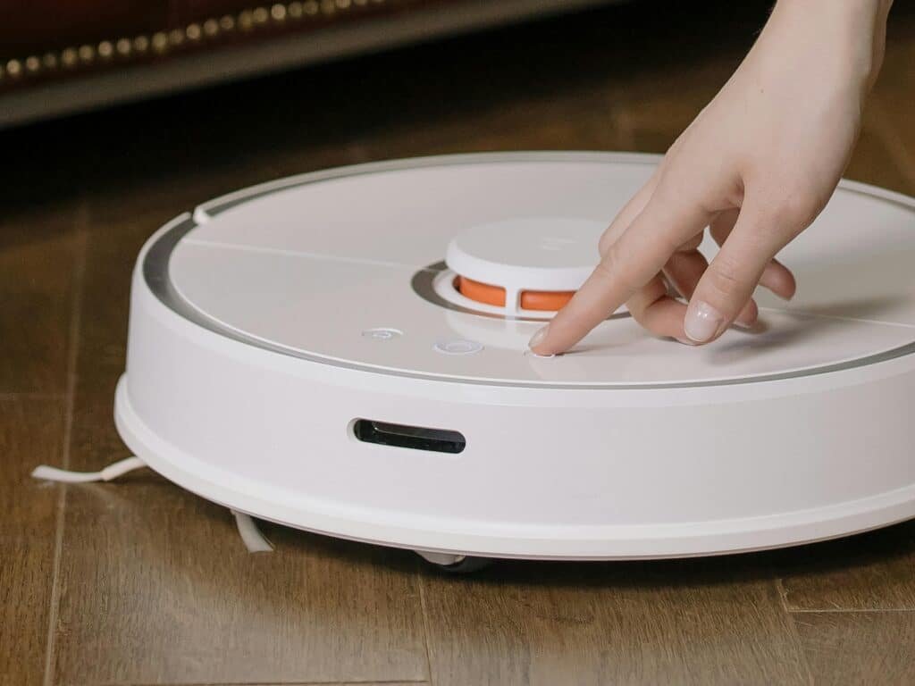 Close-up of a hand pressing a button on a robotic vacuum cleaner on a wooden floor representing How to Deep Clean Your Room.